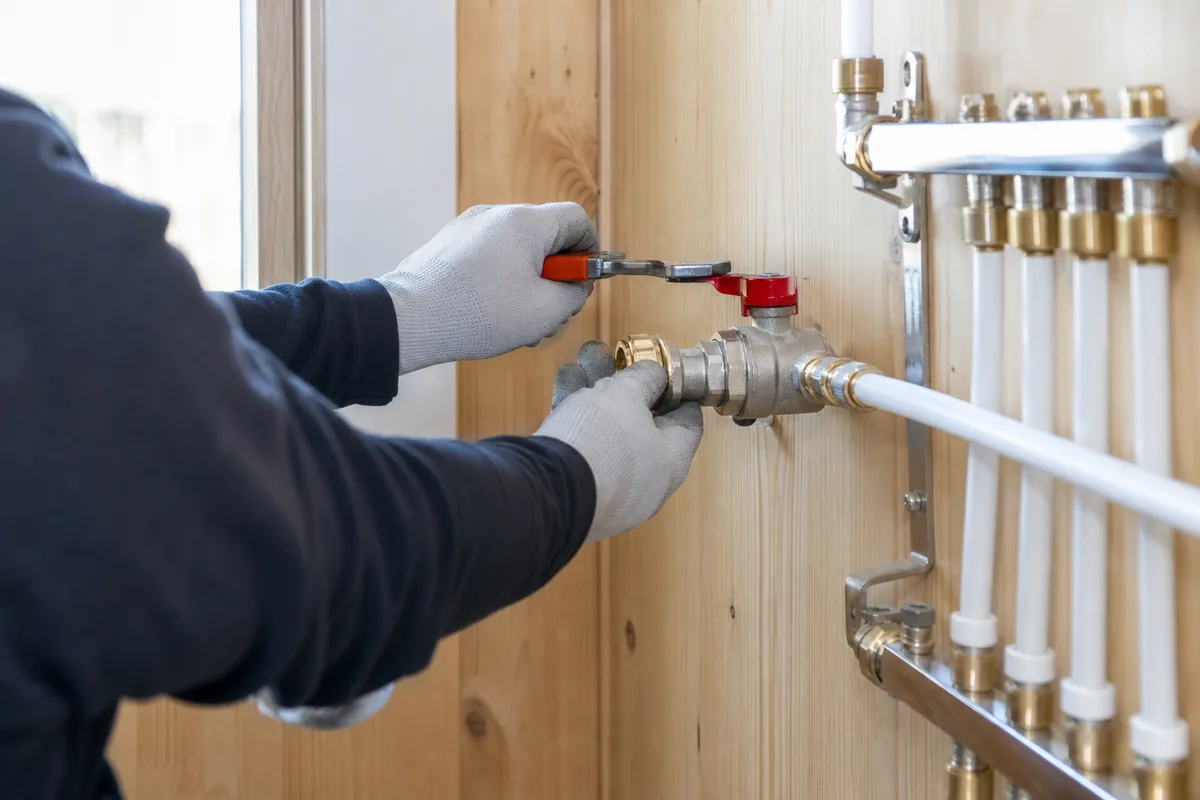 Compteur d&rsquo;eau et vanne d&rsquo;arrêt dans un local technique de maison landaise en ossature bois, lumière naturelle claire, mains gantées vérifiant un raccord