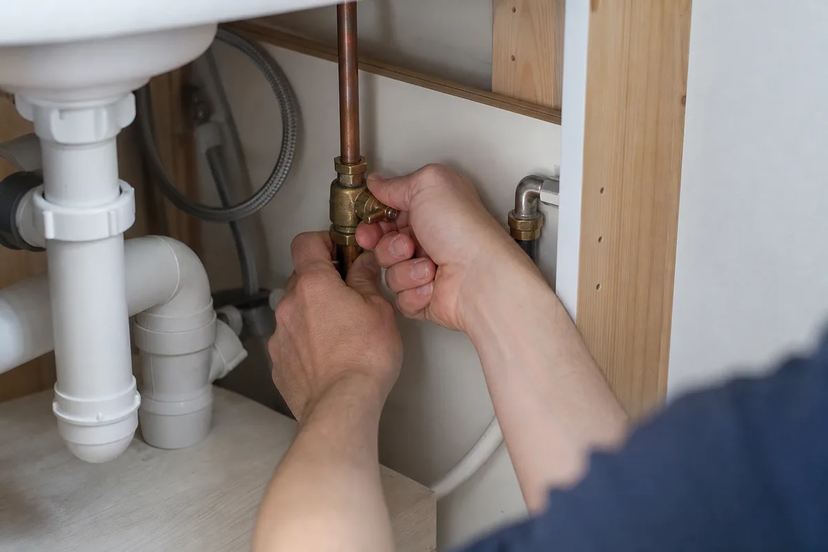 Mains fermant une vanne d&rsquo;arrêt d&rsquo;eau sous un meuble d&rsquo;évier, lumière claire d&rsquo;une maison landaise